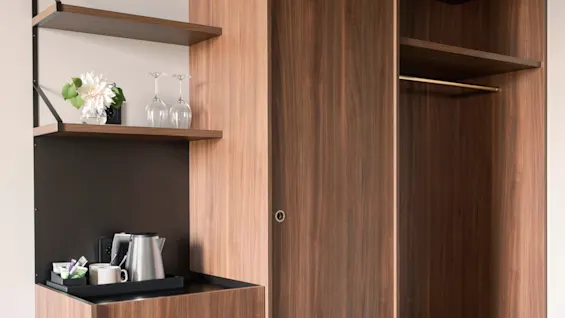 En träkök med en svart bänkskiva har en vattenkokare och muggar, bredvid öppna hyllor med glas och blommor i ett modernt, minimalistiskt rum.