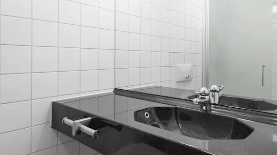 A black sink with a shiny faucet sits on a tiled countertop, reflecting in a wall mirror. Surrounding white tiled walls and a frosted glass partition complete the bathroom setting.