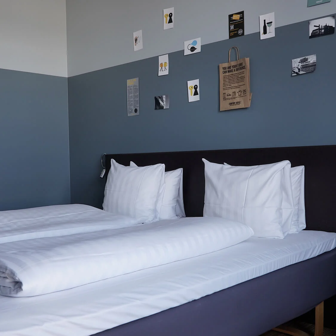 Double bed with white linens against a dual-tone blue wall, adorned with small art pieces. Text on a paper bag reads, "You and your stuff can make a difference."