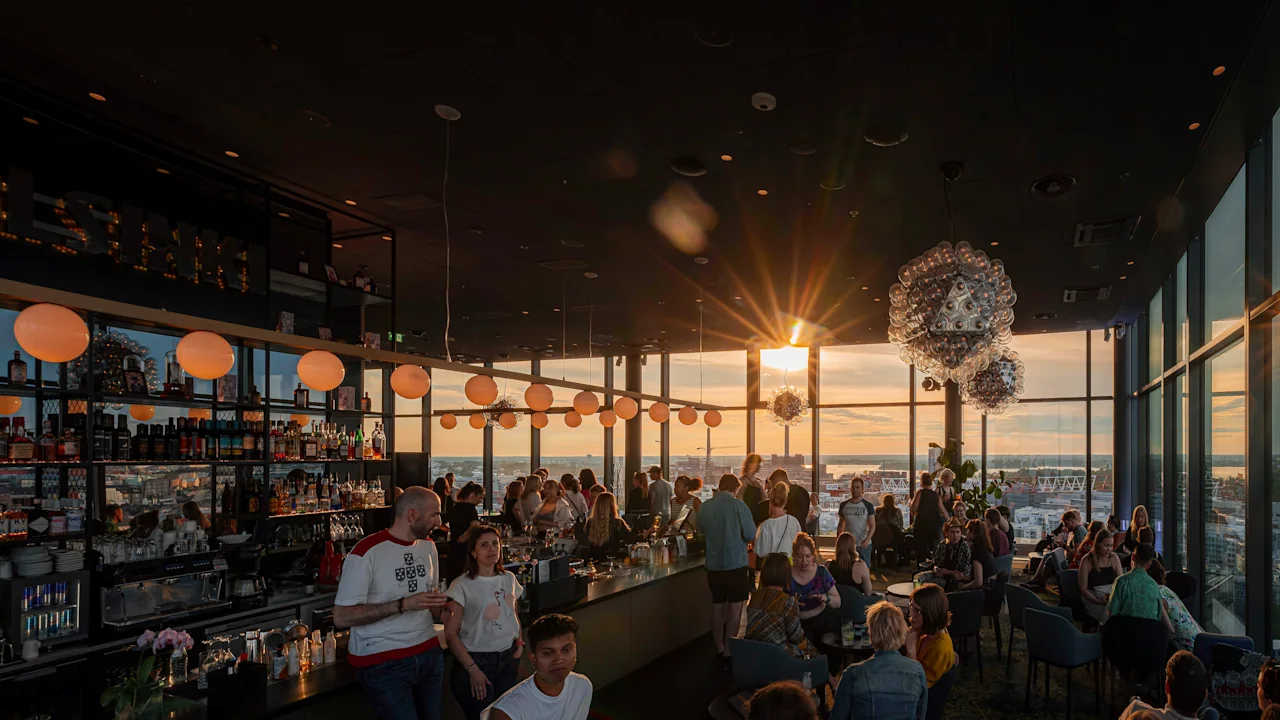 A lively rooftop bar at sunset with many people socializing. The sun shines brightly through large windows, revealing a city view. HELSINKI is visible.