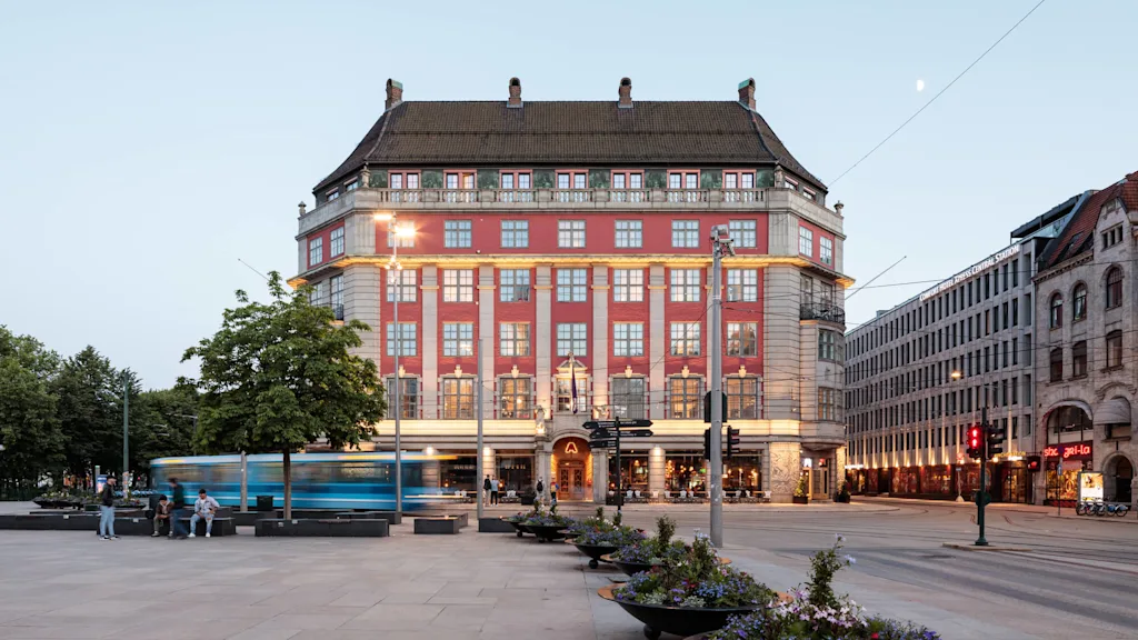 En stor, udsmykket bygning står på en plads i tusmørket, med en sporvogn der passerer. Tekst lyder: "CLARION HOTEL THE HUB OSLO CENTRAL STATION." Folk sidder i nærheden.