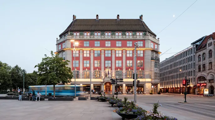 A large, ornate building stands in a city square at dusk, with a tram passing by. Text reads: "CLARION HOTEL THE HUB OSLO CENTRAL STATION." People are sitting nearby.