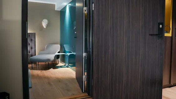 Et innbydende hotellrom med åpen dør, med en stilig sjeselong, tregulv og blått glassparti.