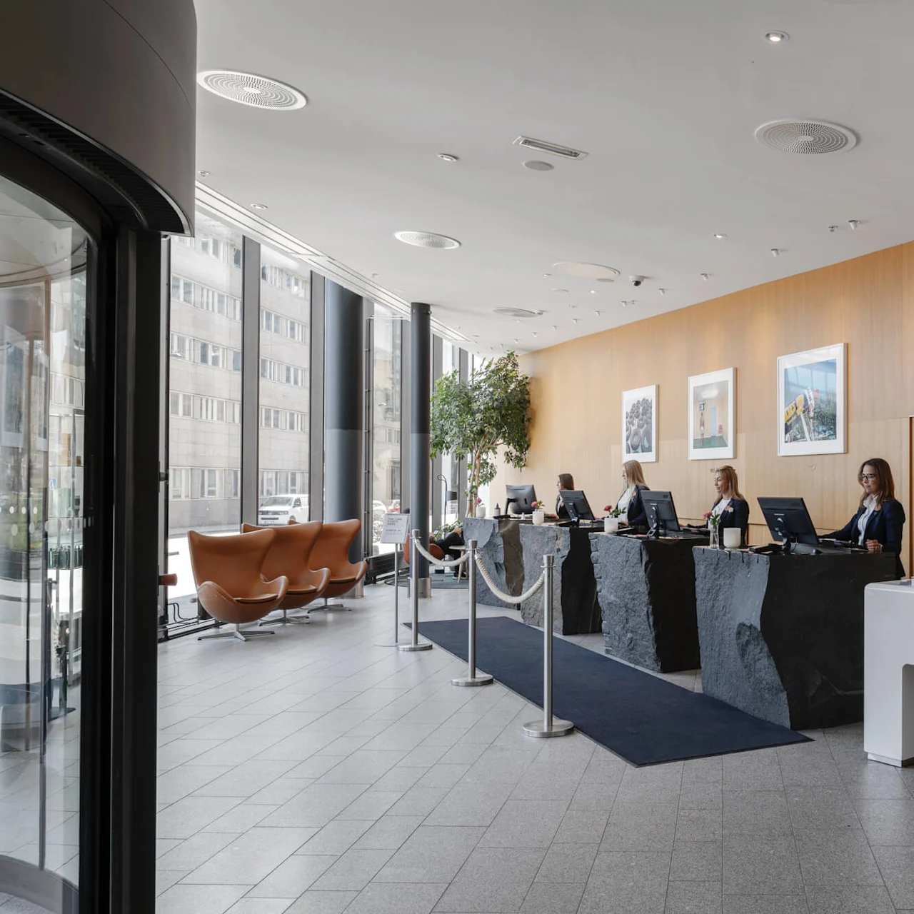 Receptionens skrivbord är bemannade av personer vid datorer i ett modernt, välbelyst rum med en trävägg och inramade bilder. Bruna stolar och stora fönster finns till vänster.