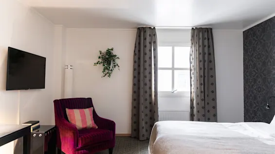 Viihtyisä hotellihuone, jossa on mukava sänky, eloisa nojatuoli, televisio ja ikkuna kuviollisilla verhoilla.