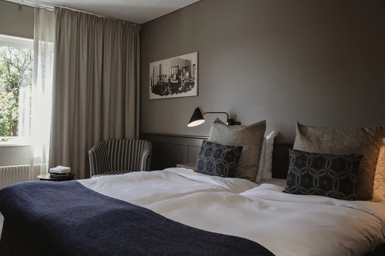 Ett mysigt hotellrum med en prydligt bäddad säng, en bekväm stol vid fönstret och en varm sänglampa.