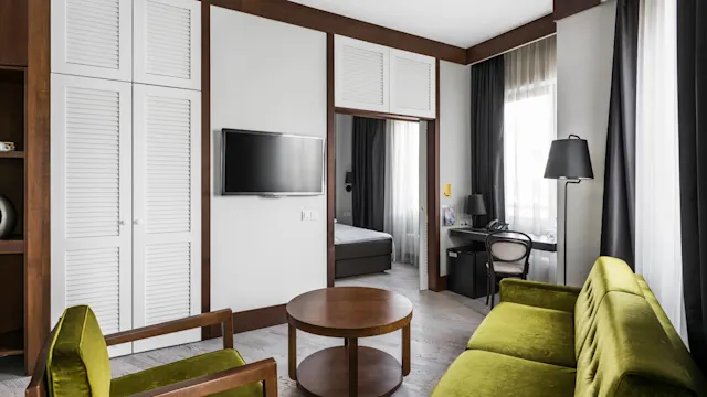 A wooden coffee table is surrounded by green sofas and chairs, placed in a modern living room with a wall-mounted TV, a large window, and a small desk area.