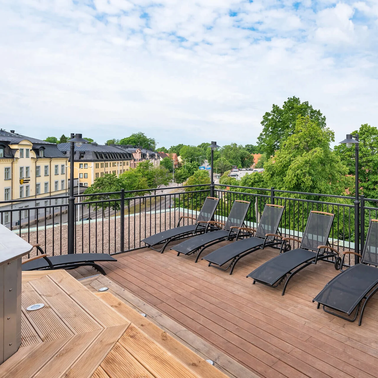 En träterrass med solstolar med utsikt över en stadssiluett med klassiska byggnader och grönska.