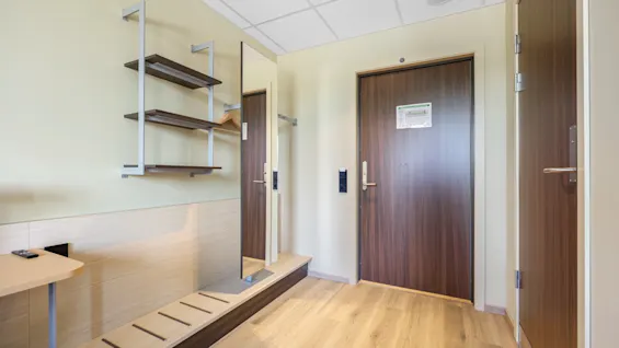 A welcoming hotel room entrance with light wooden floors, two dark doors, a wall-mounted shelf, a mirror, and a practical shoe rack.