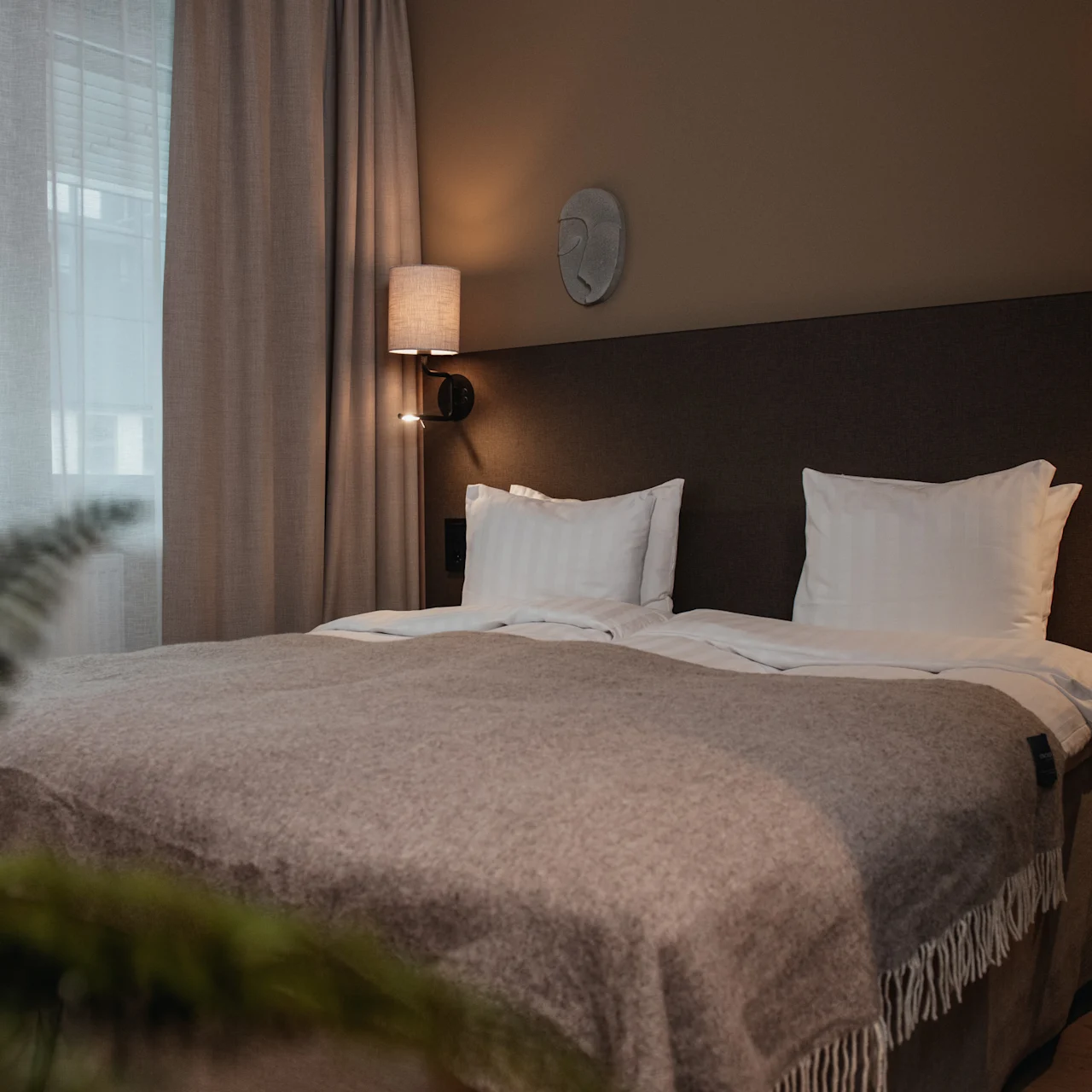 A neatly made bed with white linens and a gray blanket, flanked by two wall-mounted lamps, is in a softly-lit bedroom with curtains and a visible plant in the foreground.