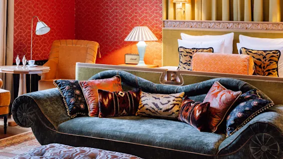 En fløyelsofa med eklektiske puter foran en seng, med bakgrunn av rødmønstret tapet, i et lyst, luksuriøst hotellrom.