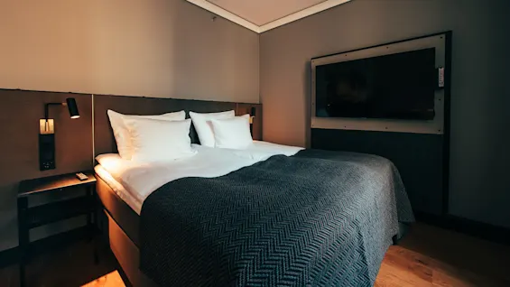 En seng med hvite puter står ved siden av et nattbord og lampe i et moderne hotellrom med veggmontert TV.