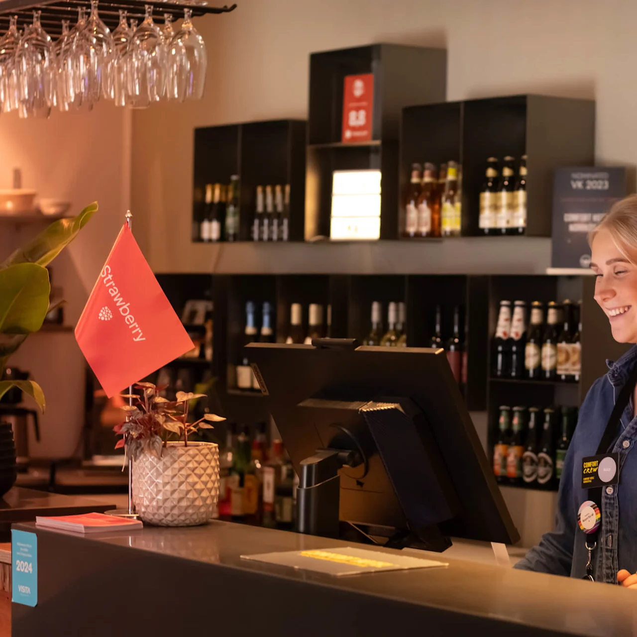 En smilende kvinne står ved en bardisk omgitt av glass og flasker, i et varmt opplyst rom med en drikkevaremeny på veggen.