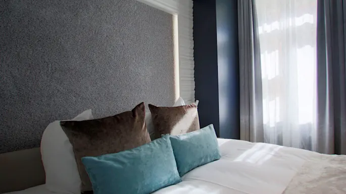 En pent oppredd seng har myke puter og et mykt teppe, plassert i et rom med teksturvegger og lys som filtreres gjennom tynne gardiner.