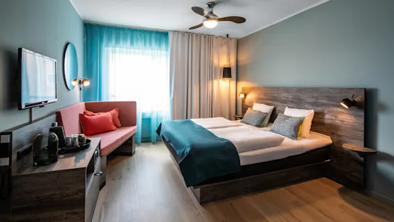 A hotel room features a double bed with teal and gray linens, a sofa, and a wall-mounted TV. Wooden floors and muted lighting create a cozy, modern atmosphere.