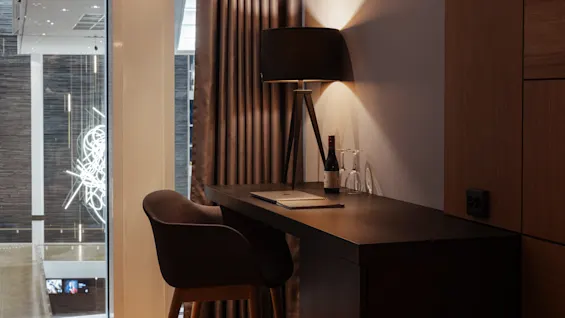 Et koselig skrivebordsområde med en lampe, flaske og glass, ved siden av et vindu med utsikt over et abstrakt opplyst rom.