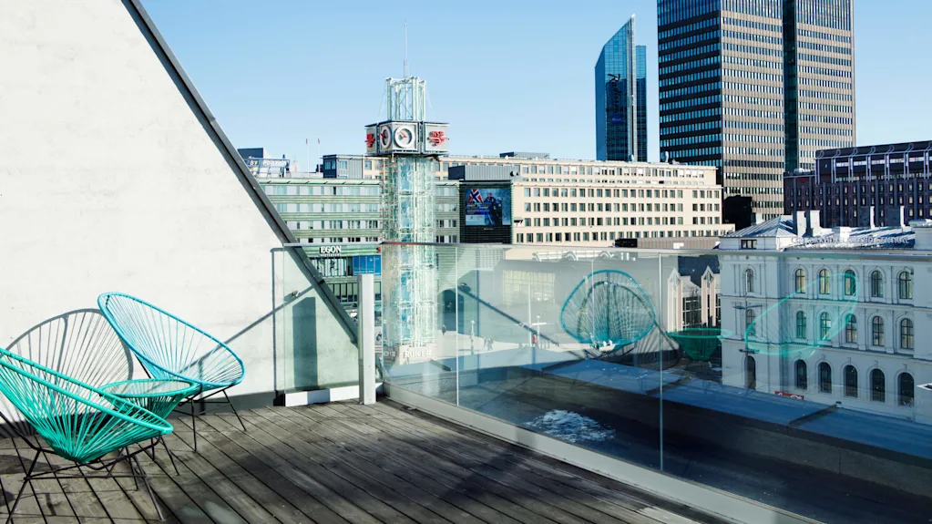 To turkise stoler står på en tretakterrasse med utsikt over moderne bybygninger, inkludert et klokketårn. Sollys kaster skygger, og glassrekkverk gir uhindret utsikt.