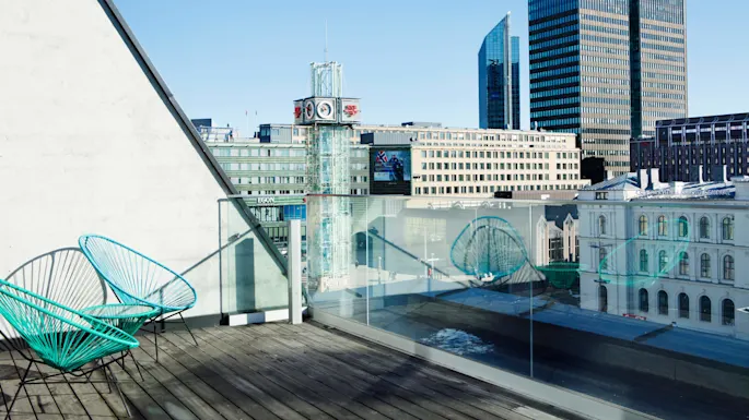 Two turquoise chairs sit on a wooden rooftop terrace overlooking modern city buildings, including a clock tower. Sunlight casts shadows, and glass railings provide an unobstructed view.