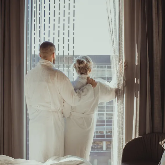 To personer i badekåper står ved et stort vindu og ser ut over byutsikten fra hotellrommet sitt.