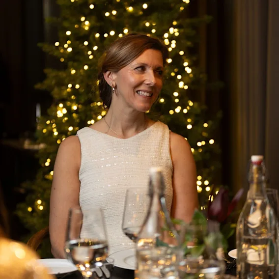 En smilende kvinne som nyter en hyggelig julemiddag med et glitrende juletre i bakgrunnen.