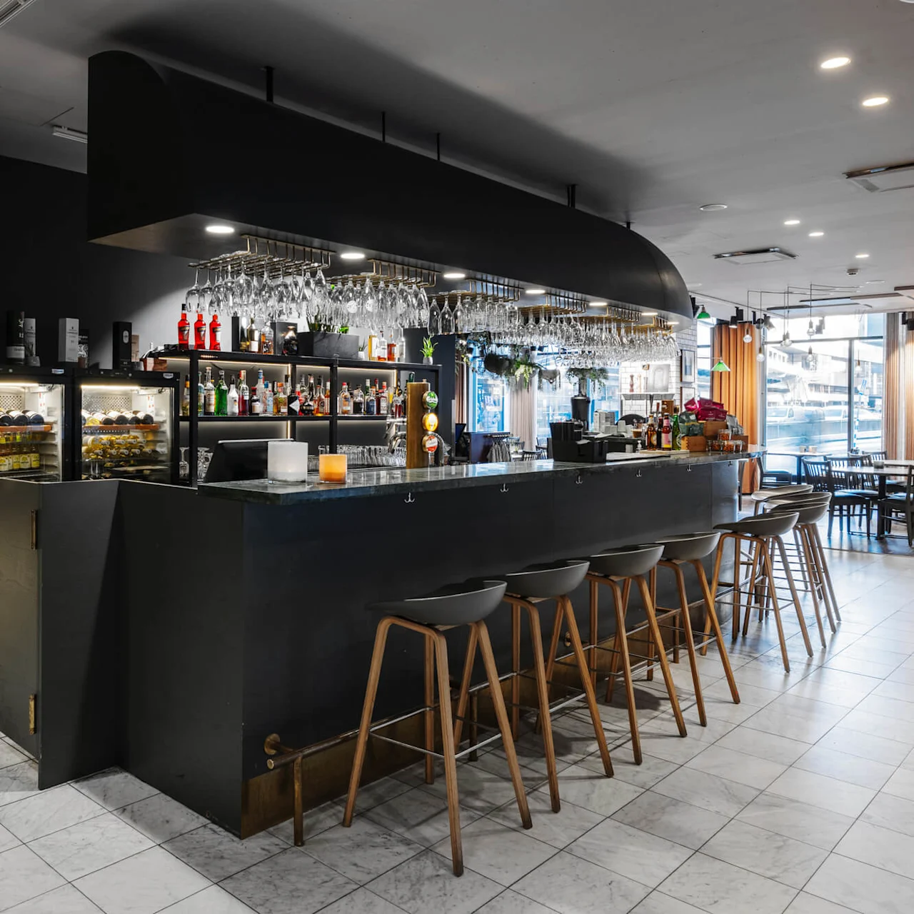 A modern bar with high stools and a black counter is stocked with glasses and bottles. Shelves display liquor, fridges hold beverages, and tables are nearby in a bright, spacious room.