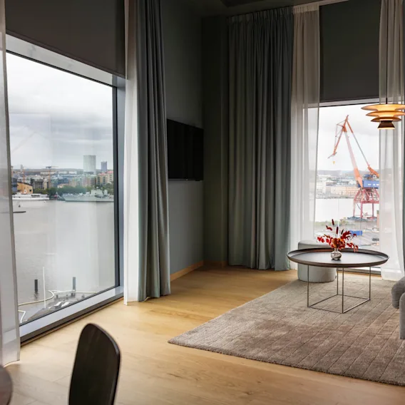 Et koselig rom med en grå sofa, et rundt bord med røde blomster og store vinduer. Utenfor er en kran synlig mot en industriell vannkant.