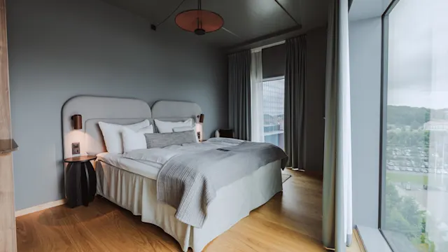 En seng med gråt og hvidt sengetøj står i et moderne hotelværelse. Store vinduer tilbyder udsigt over bybilledet. Trægulv supplerer de grå vægge.