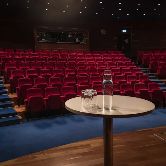Utsikt från scenen över en tom auditorium med trappade rader av röda sammetsstolar, blå matta och ett vattenbord i förgrunden.