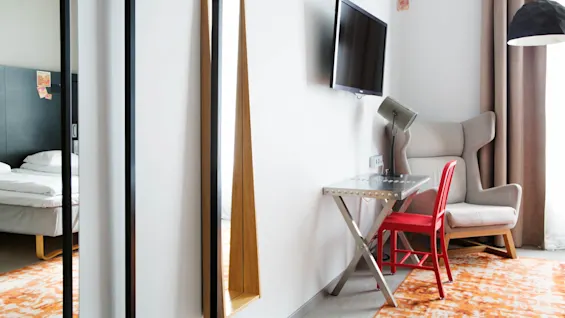 A modern hotel room features a red chair at a metal desk, facing a wall-mounted TV. A plush armchair is beside brown curtains, over an orange patterned rug.