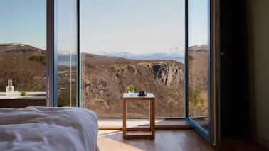 A bedroom with an open view of a mountainous landscape through large windows, a small wooden table with items beside the bed.