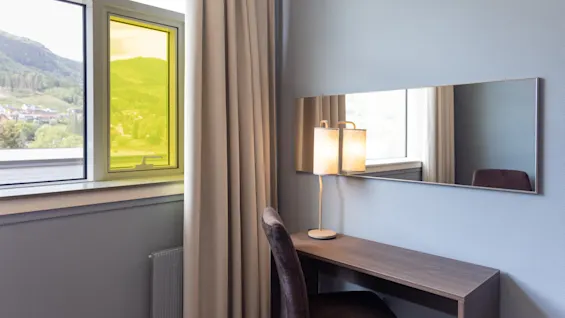 Ett litet skrivbord med en lampa och stol står vid ett fönster med gul tonad glasruta; en lång spegel reflekterar en del av utsikten och rummet, vilket förstärker den moderna, minimalistiska inredningen.
