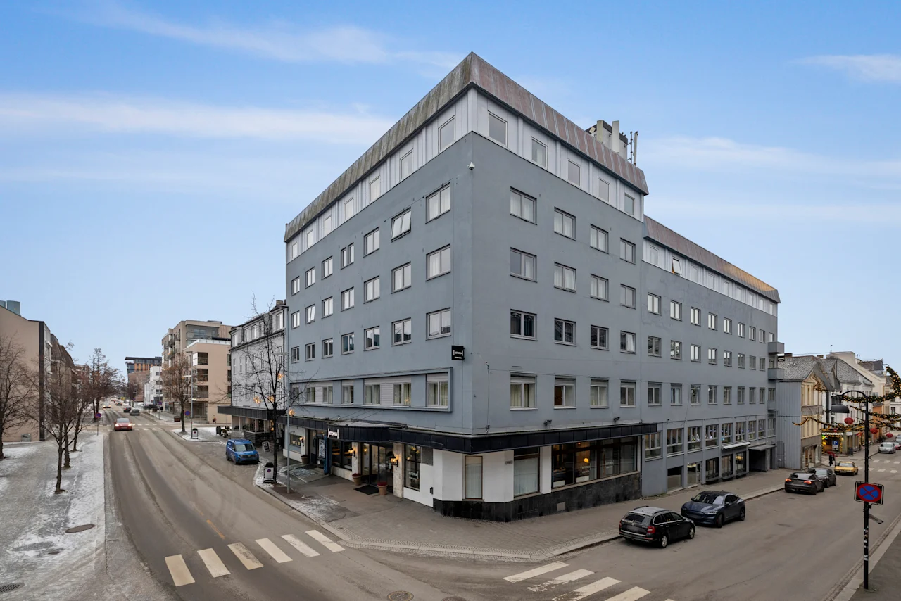 En stor, grå byggnad förankrar ett stadsgathörn, med bilar som rör sig och parkerar, och kala träd under en ljus himmel.