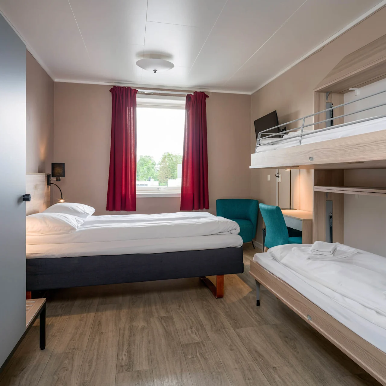A tidy hotel room features a large bed and bunk beds beneath a small desk, accented by red curtains near a window overlooking trees. A sign reads "Max 120 kg."