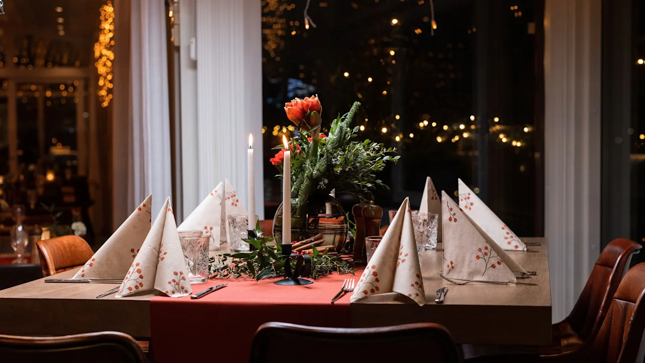 Elegant dining table, set with folded napkins and candles, surrounded by warm lights; features a central floral arrangement with red and white flowers, in a cozy restaurant setting.