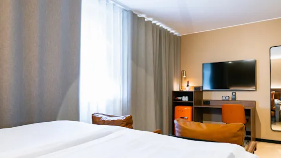 En pænt redt seng vender mod et vægmonteret TV ved siden af et skrivebord med en orange stol i et hotelværelse. Gardiner dækker store vinduer ved siden af en minibar.