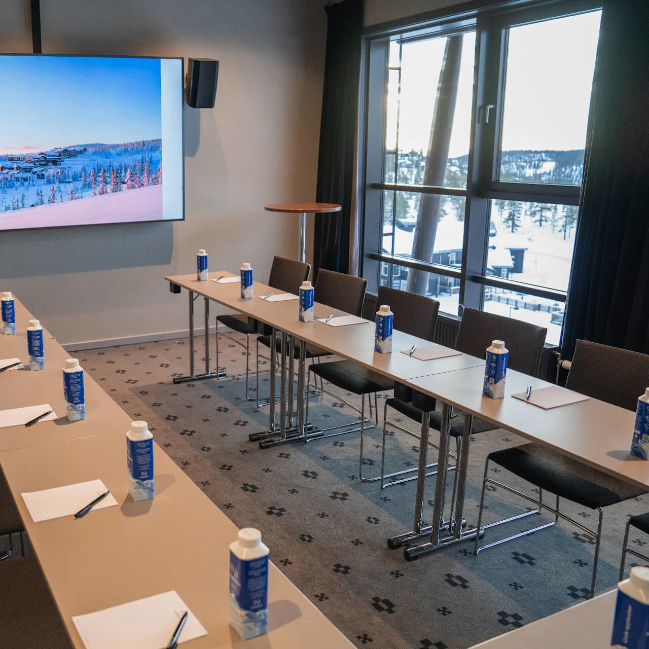 A conference room features U-shaped tables with water bottles, paper, and pens. A large screen displays a snowy landscape. Large windows offer a view of a snow-covered area.