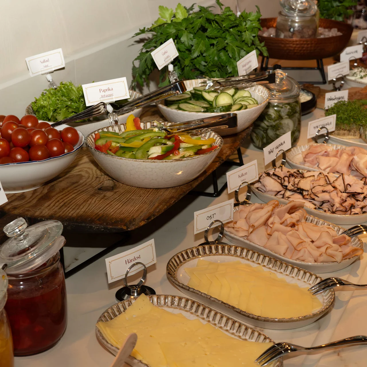 A buffet spread features various dishes including cheese, meats, salads, and vegetables, arranged with labeled name cards on a wooden surface. 
