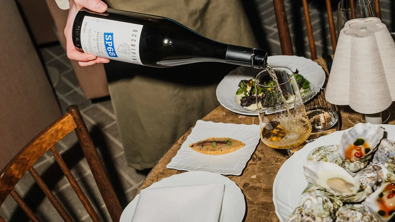 Flaske hælder vin i et glas på et spisebord med skaldyr og salat, under blød belysning.