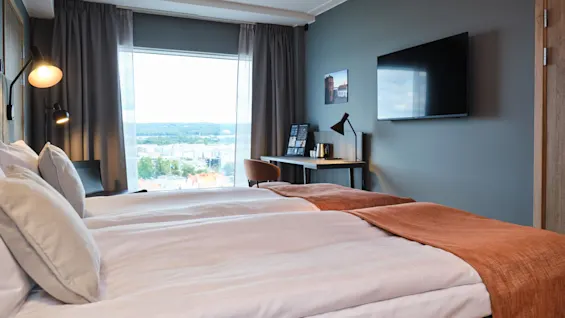En pænt redt seng med et rustfarvet tæppe står ved siden af et vindue med udsigt over bybilledet. Et skrivebord med lampe og et TV på væggen fuldender det moderne rum.