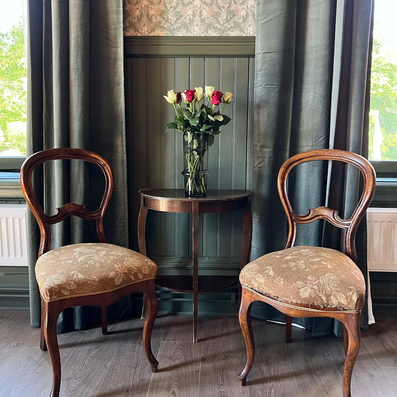 Et træbord rummer en vase med røde og hvide roser, flankeret af to vintage stole mod en baggrund af blomstrede tapet og gardiner.