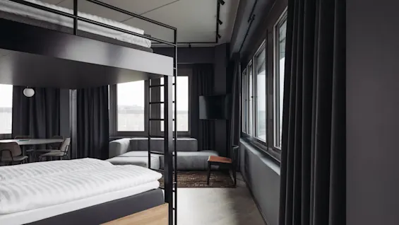 Loft bed with black frame situated in a modern room featuring a gray sofa, dining table, and large windows, creating a spacious, well-lit environment.