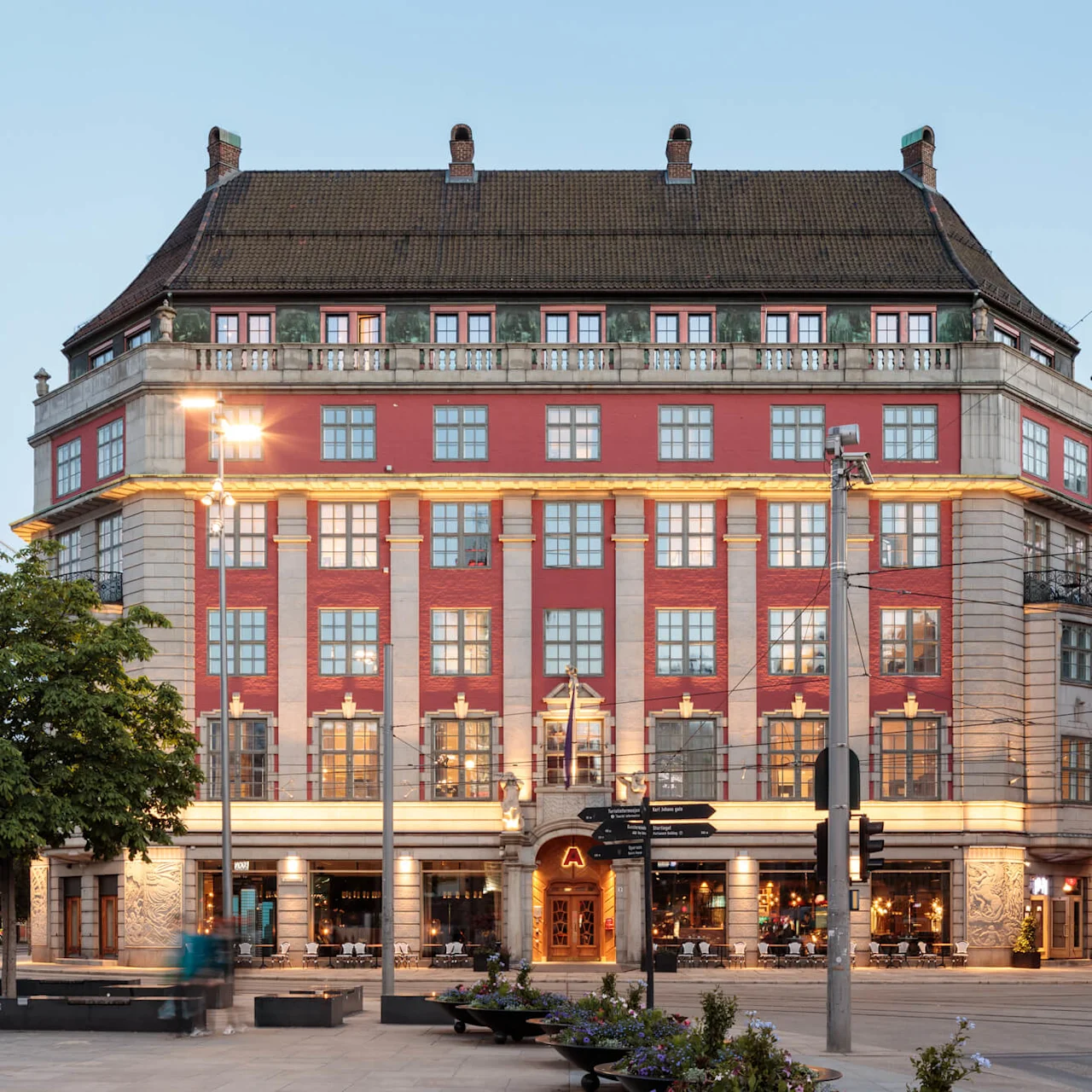 A grand, red-brick building with a steep roof stands under a clear sky. People sit on benches nearby. A sign displays "COMFORT HOTEL XPRESS CENTRAL".