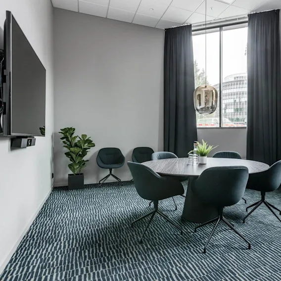 A round table with chairs sits in a modern conference room. A TV is mounted on the wall, and a potted plant decorates the space, with a large window in view. 