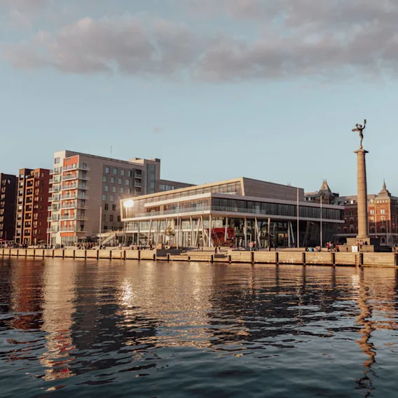 Moderna byggnader vid vattnet speglas i stilla vatten, omgivna av traditionell arkitektur och en staty på en hög kolonn. Scenen är tagen vid solnedgången med mjukt, varmt ljus.