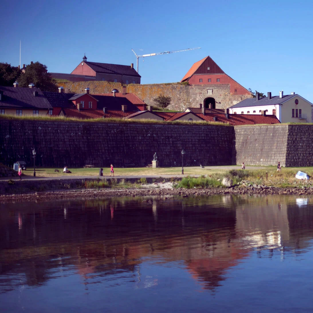 Personer njuter av en solig dag vid en lugn vattenfront med historiska byggnader och en färja i bakgrunden.