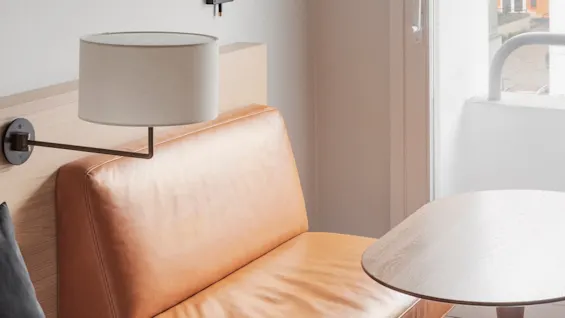 A tan leather sofa sits beside a wooden table under a black wall lamp, near a window overlooking an orange building.