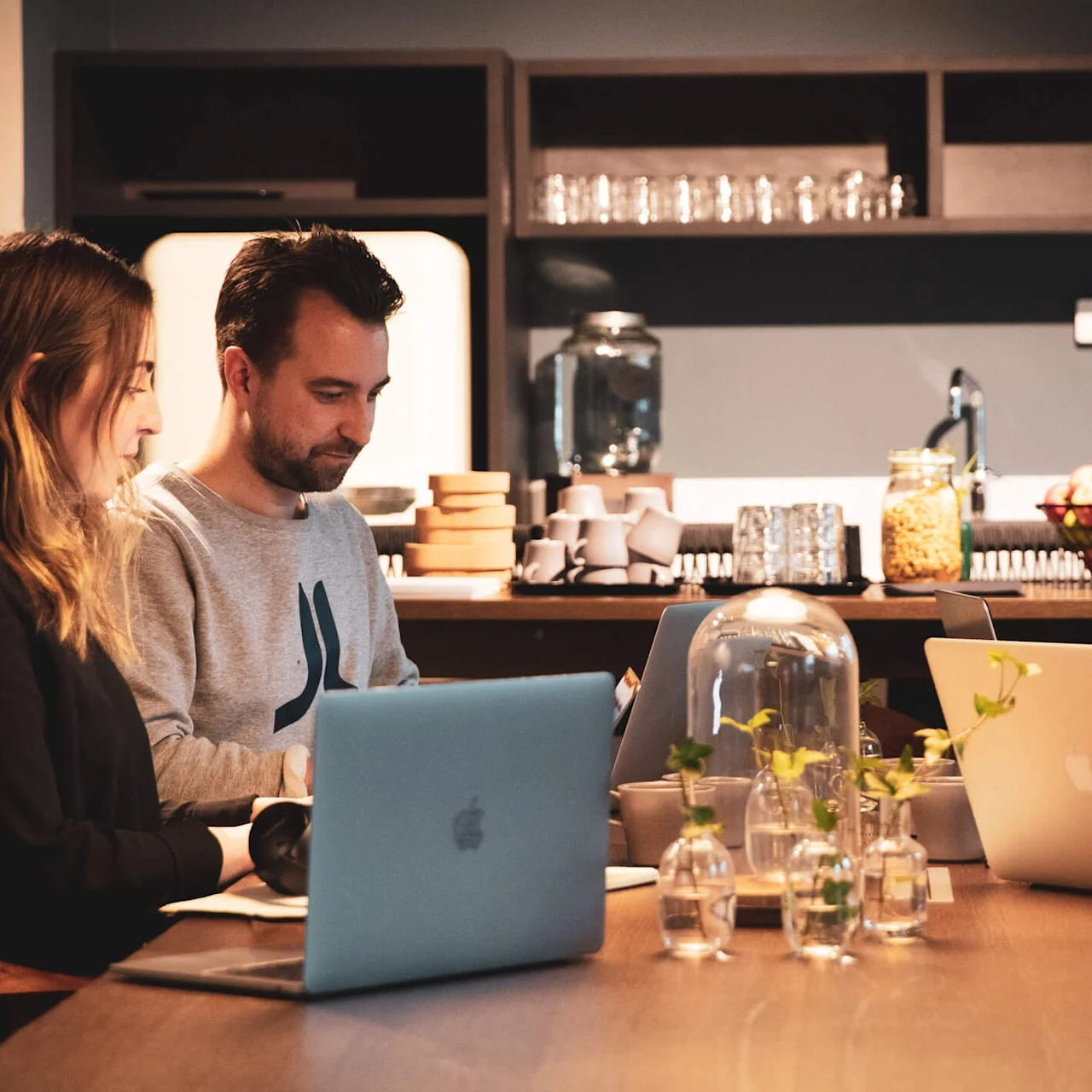 Fem personer sitter rundt et trebord og jobber med datamaskiner i et varmt belyst rom. Hyller med kopper og fat er synlige i bakgrunnen sammen med frukt og planter.