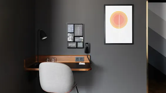 En modern arbetsplats med ett flytande skrivbord i trä, en grå stol, väggkonst, en lampa och telefon mot en mörkgrå vägg.