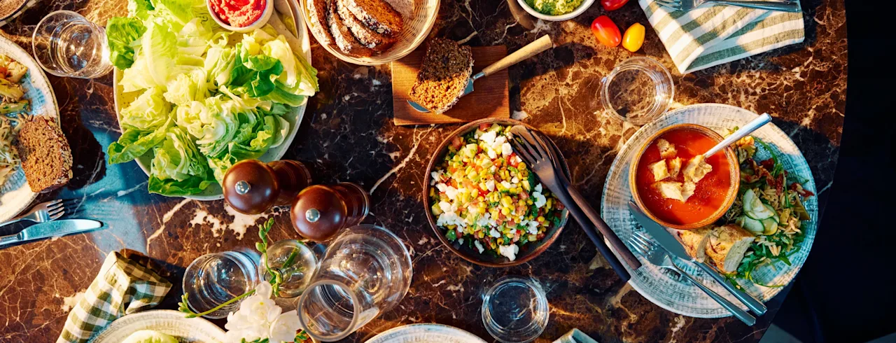 Ett marmorbord är dukat med tallrikar med olika maträtter, inklusive sallader, soppor och bröd. Glas och randiga servetter är placerade bland rätterna, vilket skapar en avslappnad matmiljö.