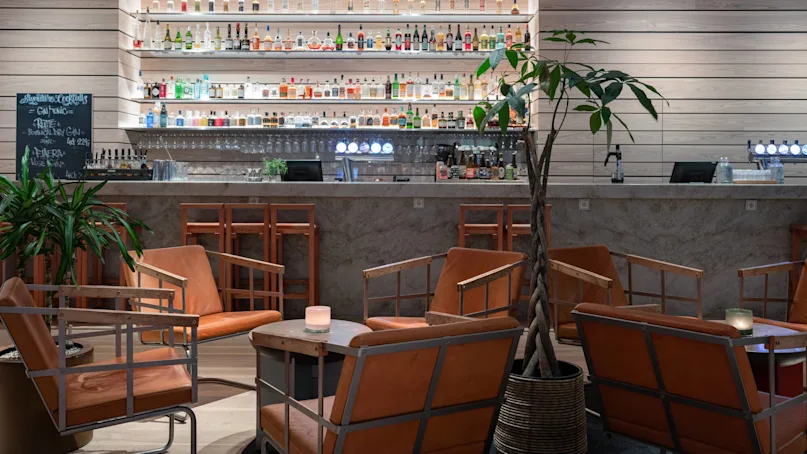 Bar with shelves of liquor bottles, providing seating with orange leather chairs around tables, surrounded by modern wood paneling and indoor plants. Chalkboard reads: "Signature Cocktails, Gin Tonic, Rattle, Aniera".  